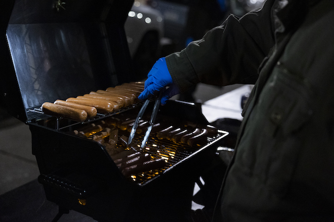 Closeup of a latex-gloved hand turning hot dogs on a grill in the dark.