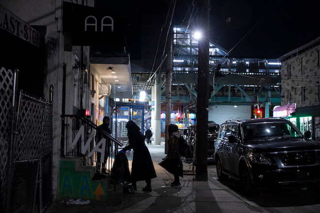 Backdropped by the Market-Frankford elevated rail line, a nighttime scene in the Kensington neighborhood of Philadelphia, where volunteer Nicole Bixler's silhouette is visible, picking up trash.