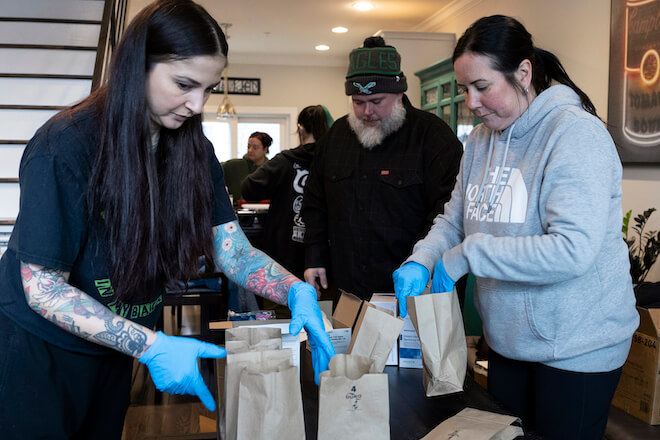 Left to right: Nicole and Wes Bixler and Kara Siegriest, three White people, stand over a table filled with brown paper bags to assemble safe-use kits in the Bixlers' Port Richmond home.