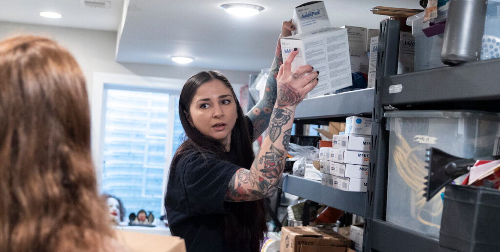 Nicole Bixler, a White woman with long black hair wearing a black top, takes a pack of sterile saline solution off a shelf in the basement of her Port Richmond home. Sterile solutions can be used for many things, including to clean out syringes and wounds.