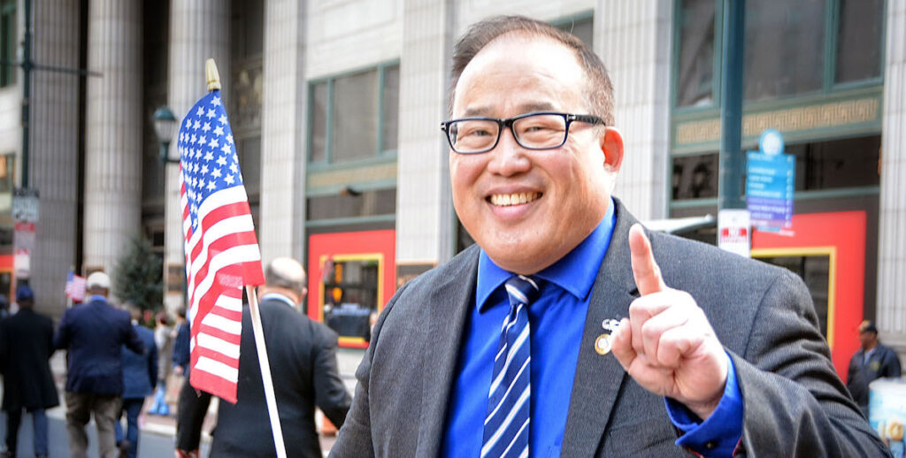 David Oh, a Korean American man wearing glasses, a suit jacket, bright blue shirt, and tie, points the index finger of his left hand in a "one" sign and holds an American flag in his right hand. He is smiling.