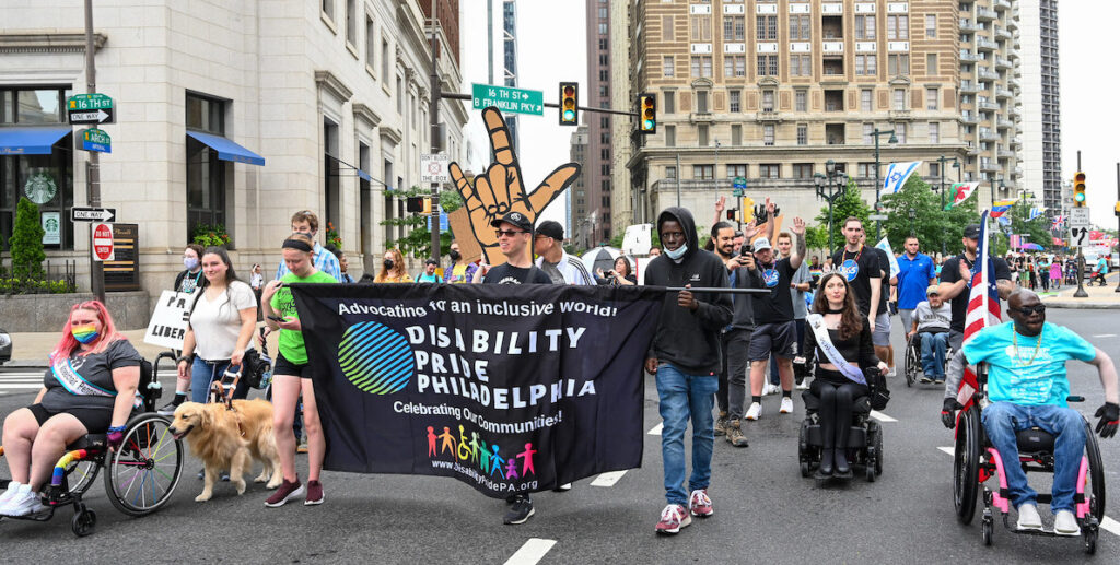A diverse group of people roll and walk along Market Street In Philadelphia. Some are in wheelchairs, another walks with a service dog, some carry a banner that says "Advocating for an inclusive world! DISABILITY PRIDE PHILADELPHIA Celebrating our communities! In the back a large sign displays a hand making the ASL sign for "love."