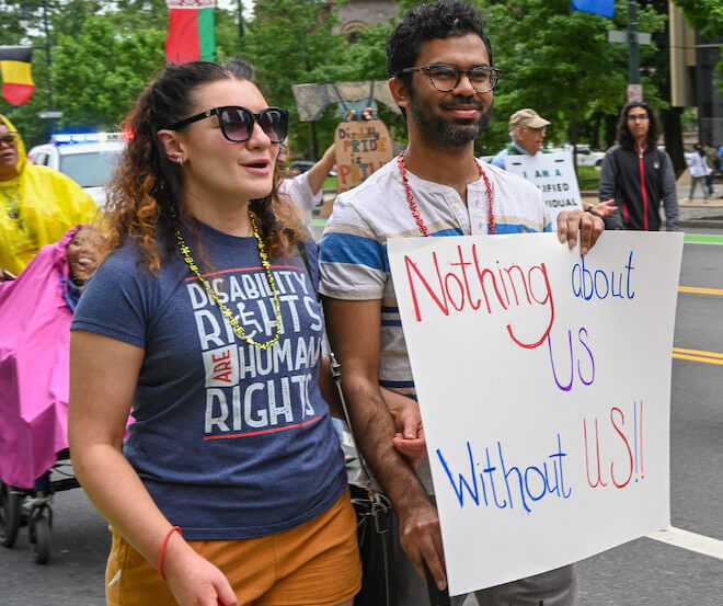 Two people walk along a street. One is a white woman with curly brown hair, sunglasses, a necklace and a blue t-shirt that reads "DISABILITY RIGHTS ARE HUMAN RIGHTS." She is holding the arm of a brown man with curly black hair and glasses. He is wearing a white shirt with strikes and holding a sign that says, "Nothing about US Without US!!"