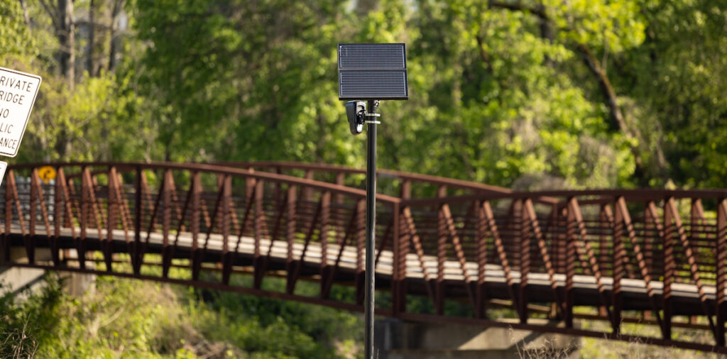 An automatic license plate reader (ALPR) from Flock Safety stands in front of a pedestrian bridge and wooded area.