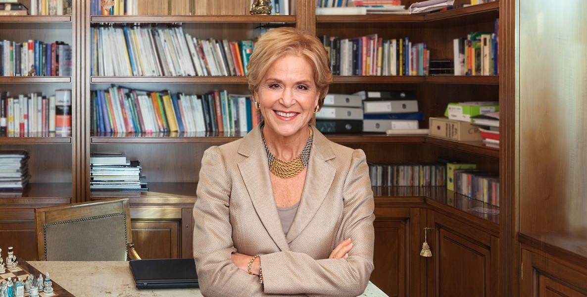Judith Rodin, a White woman, sits on an office desk in front of full bookshelves. She's wearing a beige silk suit jacket and large gold and silver necklace. She's the newest member of Hilco's board, the company behind the Bellwether District.