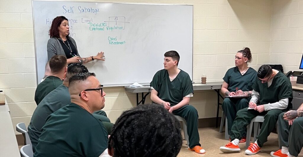 People incarcerated at the Denver Downtown Jail wearing dark green shirts and pants and bright orange shoes, sit around a room looking at a woman with dark hair who is standing by a whiteboard with the words "Self Sabotage" written on it. These are participants in a therapy session for people undergoing medically assisted treatment for addiction.