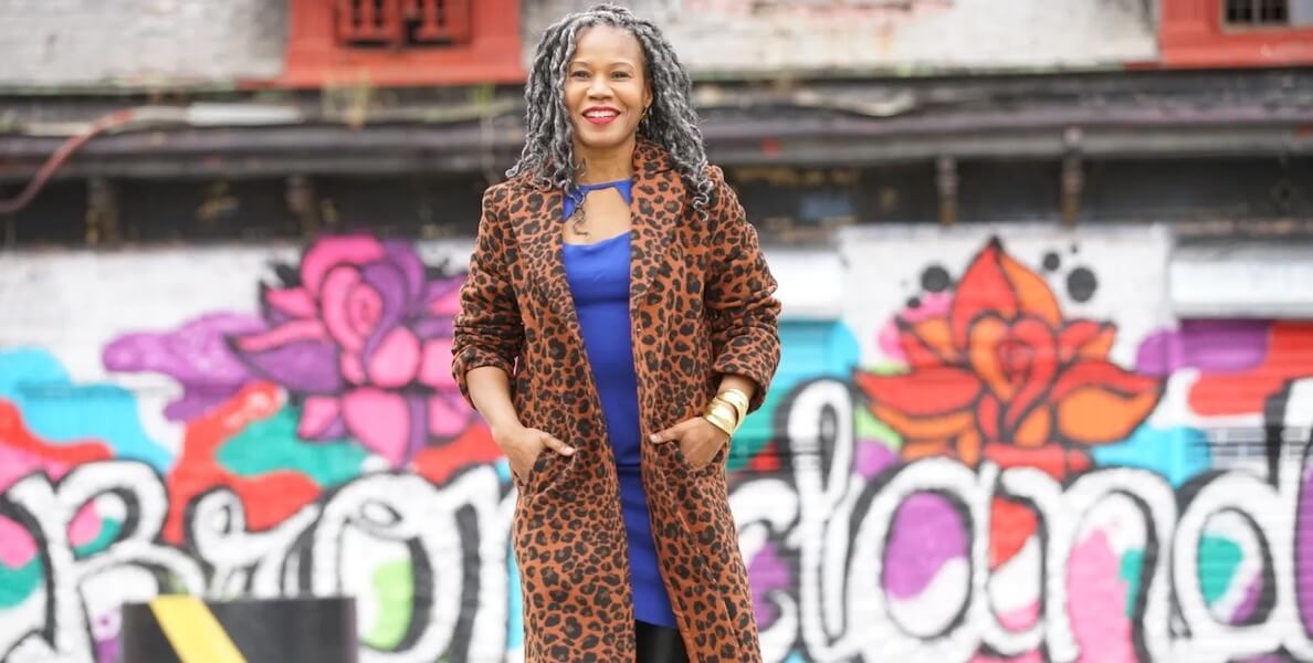 Majora Carter, a Black woman with grey curly hair past her shoulders, wearing a purple dress and animal-print coat, stands before a wall with a colorful, graffiti-style mural that says "Bronxland."