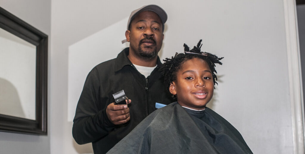 Barber Mal Robinson, a Black man with a tight beard, grey cap and black collared jacket, holds clippers while standing behind a Black child wearing a barber's cape and getting a shape up.