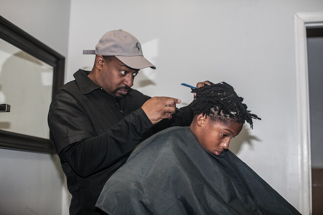 Barber Mal Robinson, a Black man with a tight beard, grey cap and black collared jacket, holds shapes up the back of a hairstyle on a Black child in a barber's cape.