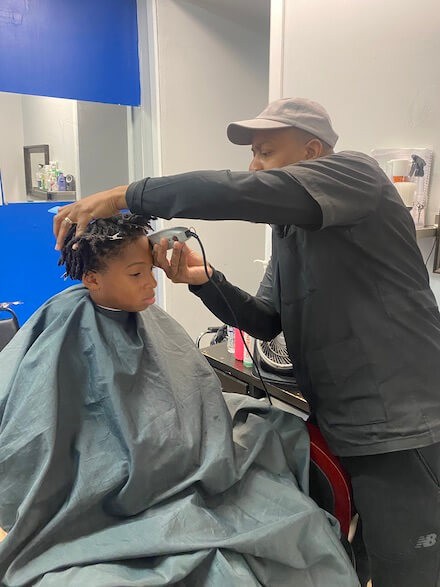 Barber Mal Robinson, a Black man in a grey cap, long-sleeved black shirt, and grey shirt overtop, holds clippers to the head of a Black child wearing a barber's cape and getting a shape up.