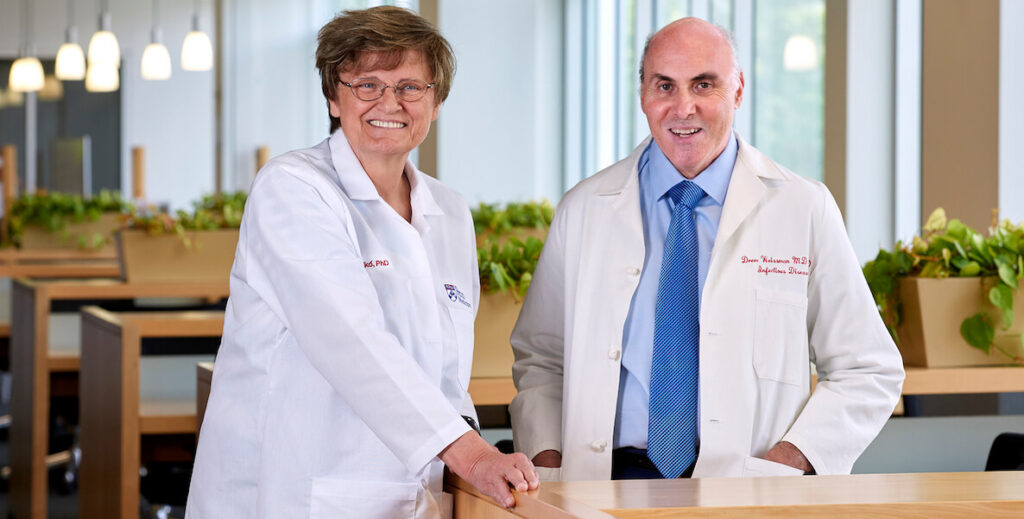 Dr Katalin Karikó, a white woman with short brown hair and glasses in a medical coat, and Dr Drew Weissman, a white man with a bald head wearing a doctor's jacket, blue collared shirt and tie, stand together at Penn.