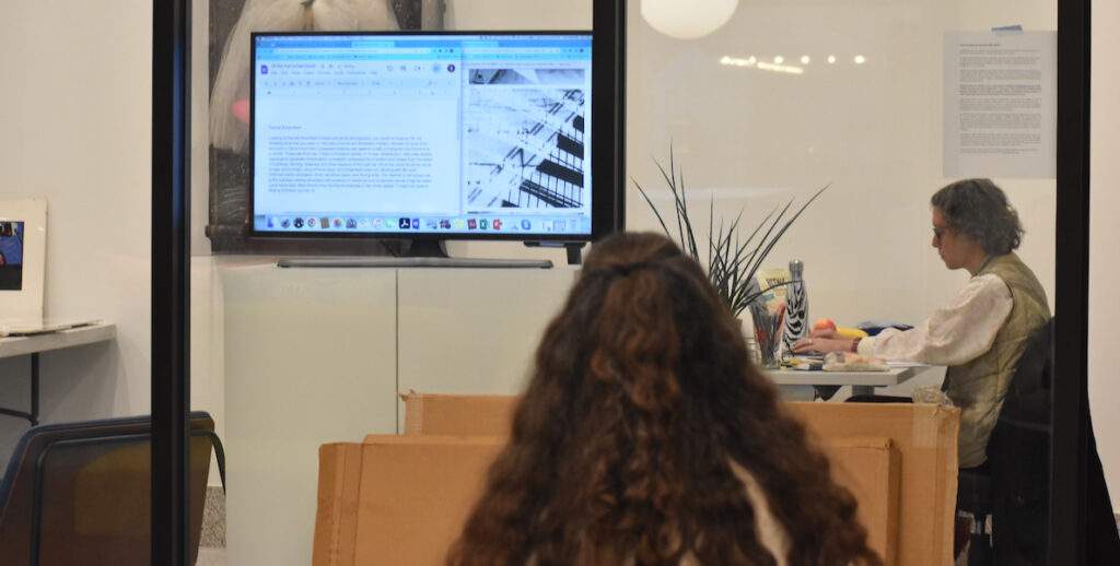 The back of a person with long brown hair is shown. The person's body is facing a window into an office at Gershman Hall, where 60 wrd/min art critic Lori Waxman is typing on a computer.
