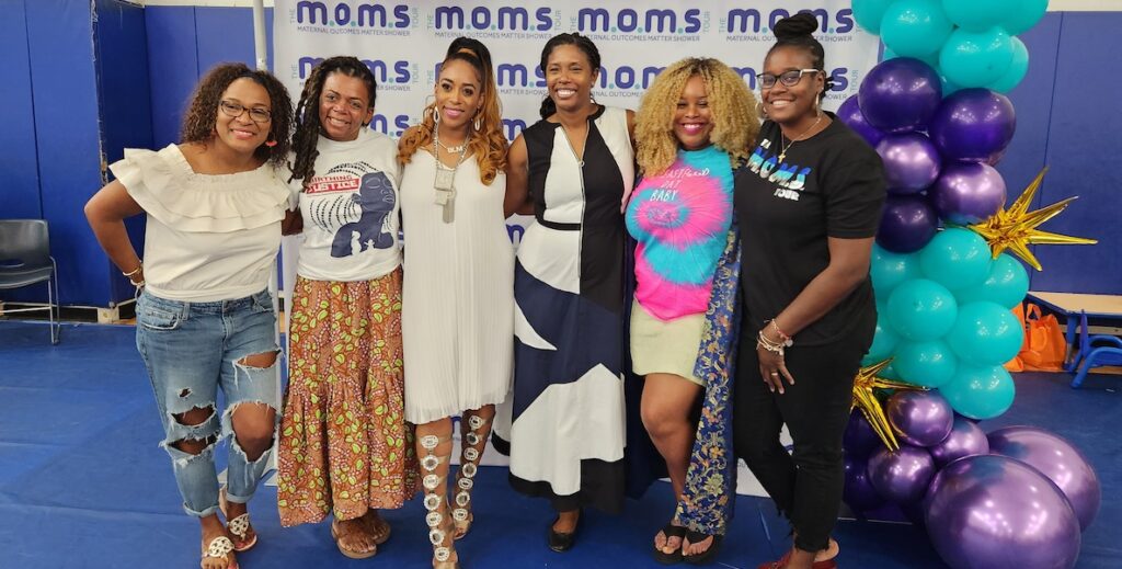 A line of well-dressed Black women stand before a backdrop that says M.O.M.s at a Philadelphia stop on the national M.O.M.s (Maternal Outcomes Matter) tour.