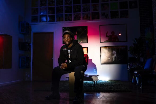 A portrait of CJ Wolfe, a Black man in a black outfit, somewhat seated in his studio, and lit by a colorful spotlight. Wolfe is the founder of the photography studio Immortal Vision in Kensington in Philadelphia.