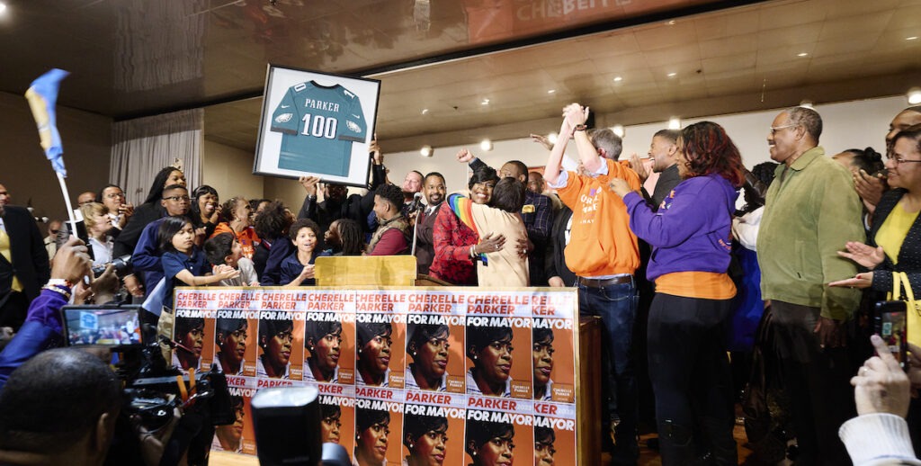 Cherelle Parker (red flower patterned top), Mayor-elect presented with an Eagles "Parker 100" jersey, hugging mentor Marian Tasco (gold necklace, rainbow sweater), former City Councilmember; Aren Platt (white, beard, glasses, orange CP hoodie), Senior Advisor to Cherelle Parker; Sinceré Harris (purple CP hoodie), Campaign Manager Thousands gathered at the Sheet Metal Workers Local 19 at Columbus Blvd and Washington Ave to celebrate the 100th Mayor of Philadelphia: Cherelle Parker. November 7, 2023 for The Philadelphia Citizen