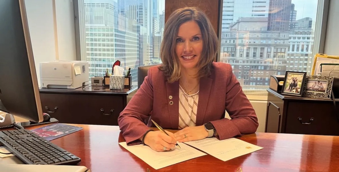 Christy Brady, a CPA from the City Controller's office, sits at her desk and smiles. She is a white woman with shoulder-length hair. She is wearing a raspberry jacket and white blouse and appears to be writing on a piece of paper. Behind her: the skyline of Center City, Philadelphia.
