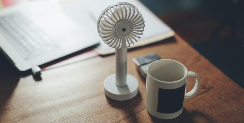 Photo of a small upright desk fan, empty mug and paperwork on top of a wooden desk to represent menopause at work.