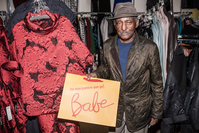 Ted Hall, the 87-year-old proprietor of Babe, a women's boutique at 110 S. 52nd Street is one of the last Black business owners along a historically Black business corridor. He stands, wearing an olive driving jacket and grey hat, next to a bright red floral pantsuit, holding a bag that says: "for everywhere you go! Babe"