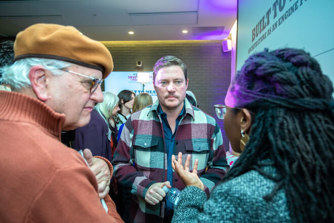 Architect Michael Murphy, a white man with light brown hair and a mustache wearing a plaid button-down jacket, speaks with a White mane with white and grey hair, glasses, an orange sweater and an orange beret and a Black women with braided locks, glasses, and a black and white tweet coat.
