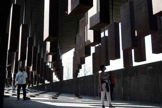 Two people walk beneath large, beams representing lynchings that hang from the ceiling of the Monument for Peace and Justice in Montgomery, Alabama, designed by MASS Design Group and architect Michael Murphy.