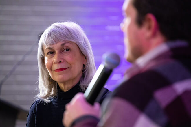 Architecture critic Inga Saffron, a White women with shoulder-length white and grey hair wearing red lipstick and a black turtleneck, looks at architect Michael Murphy (back to us), who is speaking into a microphone.