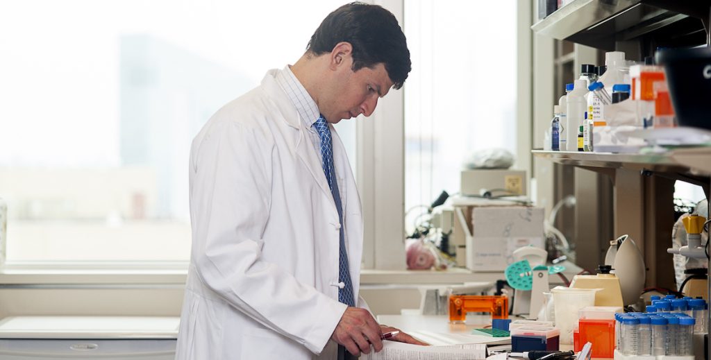 Researcher and M.D. David Fajgenbaum, a White man in a doctor's coat, stands, looking at a paper on a countertop full of medical and research equipment.