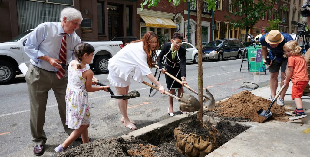 Paul Levy, a White man with white hair wearing a collar shirt, tie, pleated khaki pants and dress shoes, and a child with pigtails and a dress, hold a shovel of soil destined for a new tree alongside a city street in Philadelphia. Other people are shoveling dirt too.