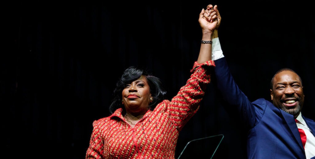 Philadelphia Mayor Cherelle Parker, a Black woman with shoulder-length black hair in a red and gold dress holds the raised hand of Kenyatta Johnson, City Council President, a Black man with a short beard wearing a blue suit, white shirt and red tie, and smiling.