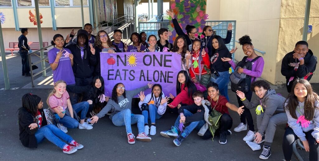 Students outside a school stand and sit. Some hold a large purple sign that reads "NO ONE EATS ALONE."