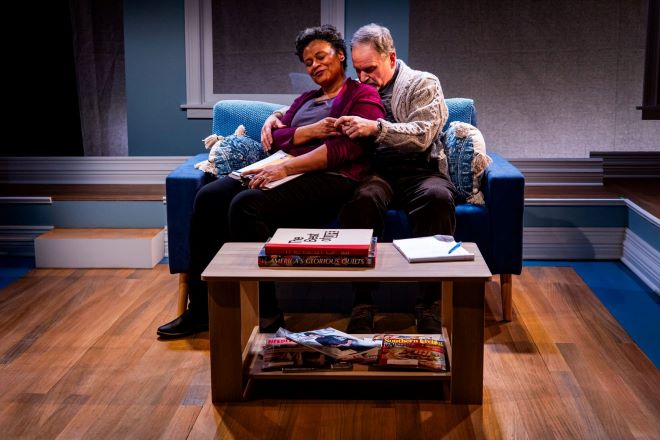 Actors Melanye Finister and David Ingram sit together on a couch in a scene from the play <i>Ladysitting</i>