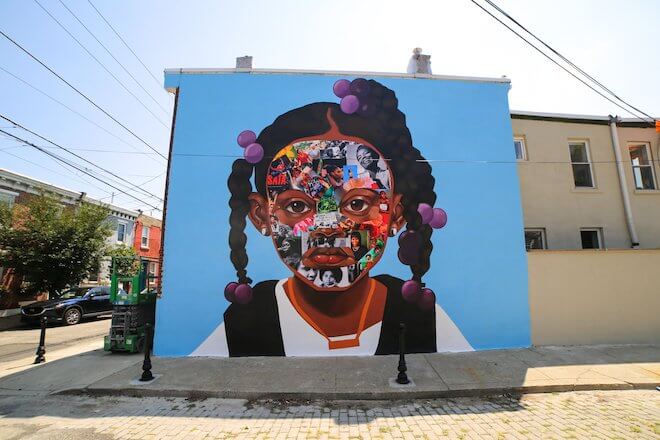 Monée. Photo by Conrad Benner of Streets Dept. A mural on the side of a building with a sky blue background and the face of a young Black girl with her hair in three twists, across her face is a collage. By artist Nazeer Sabree.
