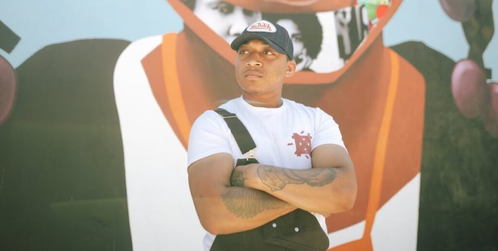 Artist Nazeer Sabree, a Black man wearing a black baseball cap, black overalls with one strap not hooked and a white t-shirt stands in front of one of his murals. Photo by Guarida.