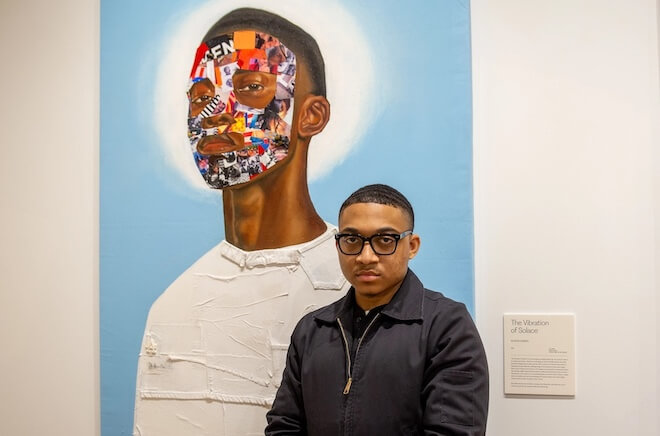 Artist Nazeer Sabree, a Black man with close-cropped hair looking serious and wearing black-framed glasses and a zip-up black jacket stands in front of