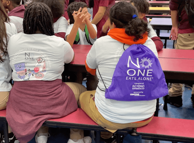 Students sit at a cafeteria lunch table. Two students sit their backs to us. One, a girl with black braids, wears a t-shirt that says "No One Eats Alone." To her right, a students in a white t-shirt over an orange hoodie with a curly black ponytail wears a backpack that says "NO ONE EATS ALONE."
