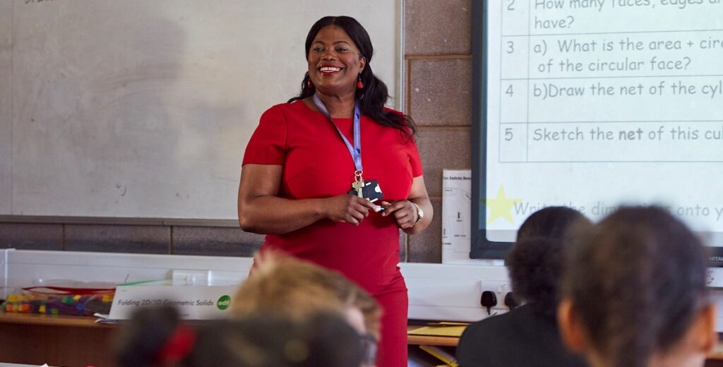 A Now Teach teacher, a Black woman in a red dress smiles while standing next to a wipe board at the front of a classroom of children.