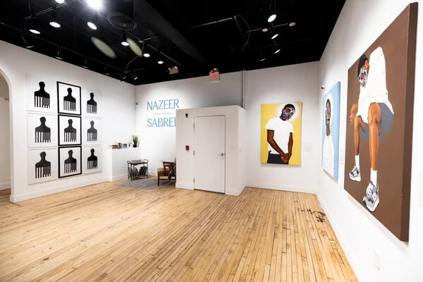 Inside an art gallery with art of a hair pick, portraits on white walls and a wooden floor, part of the Pursuit of Healing exhibit of work by Nazeer Sabree at Paradigm Gallery, courtesy of the gallery.