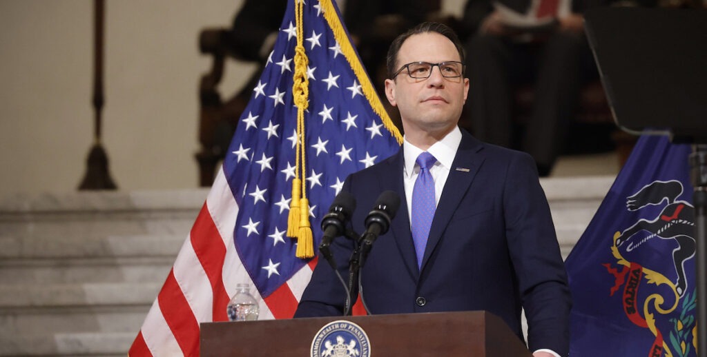 PA Governor Josh Shapiro stands before an American flag and behind a podium. He is a white man with short brown hair wearing a navy suit, white shirt and periwinkle tie.