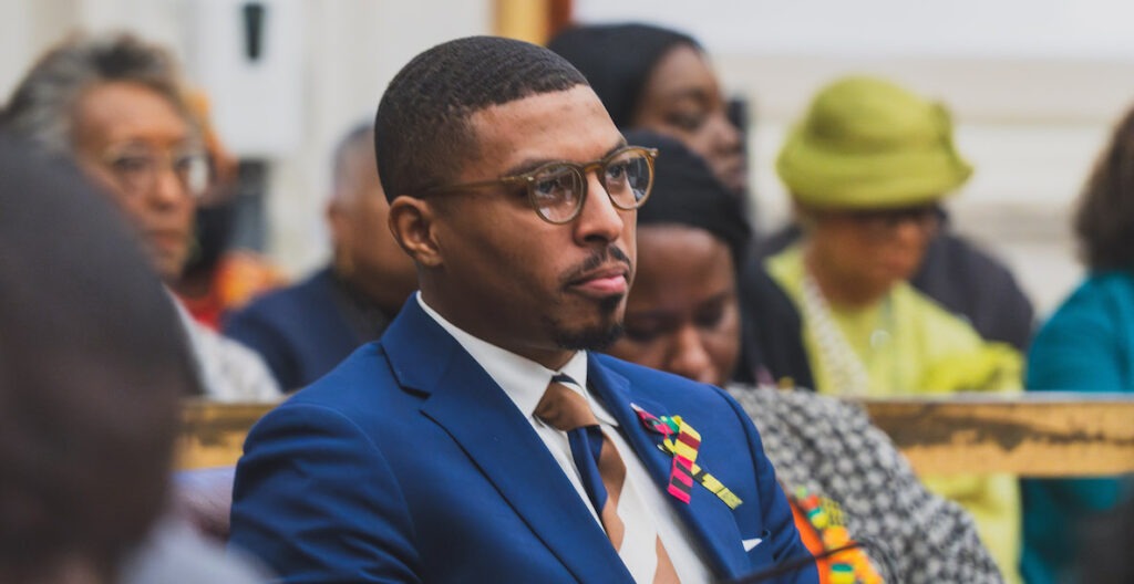 Nicolas O'Rourke, At-Large member of Philadelphia City Council, is seated in Council chambers. he is a 30-something Black man with closely cut hair wearing tortoise shell framed glasses, a blue suit jacket, white shirt and striped tie, along with an African ribbon on his lapel.