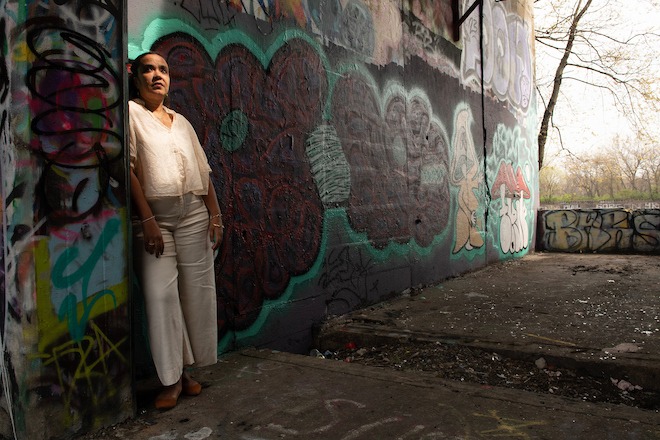 A dim portrait of Adjoa Jones de Almeida, the new director of Forman Arts Initiative, a Latina with her hair pulled back, wearing a white blouse and pants. She stands against one wall of Graffiti Pier in Philadelphia. Photo by Sabina Louise Pierce.