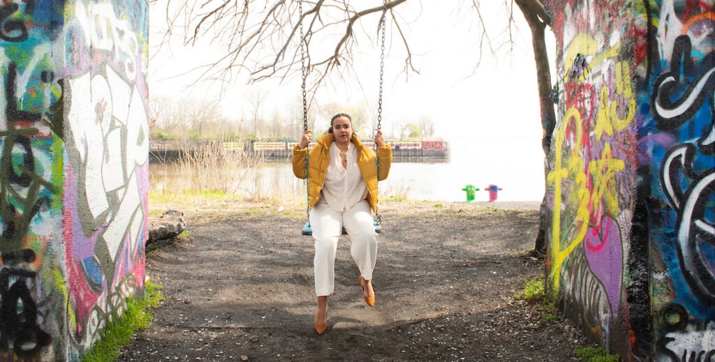 Adjoa Jones de Almeida, a Black Latina woman with her hair pulled back, sits on a swing at Graffiti Pier in Philadelphia. She is wearing a white blouses and pants, with mustard yellow shoes and jacket. On either side of her are graffitied walls. Behind her is a small patch of the Schuylkill River.