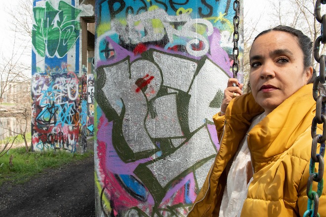 A partial portrait of Adjoa Jones de Almeida, the new director of Forman Arts, Initiative, a Latina with her hair pulled back, wearing a puffy mustard yellow jacket and white blouse. She grips with one hand the chain of a swing she's seated on (not pictured). Behind her stands two walls of Graffiti Pier in Philadelphia. Photo by Sabina Louise Pierce.