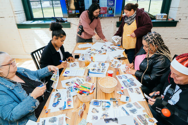 Six people sit and stand around a table, cutting photos and pasting them on white sheets of paper. They are making expressions of their wishes for things the City of Philadelphia should include in the budget.