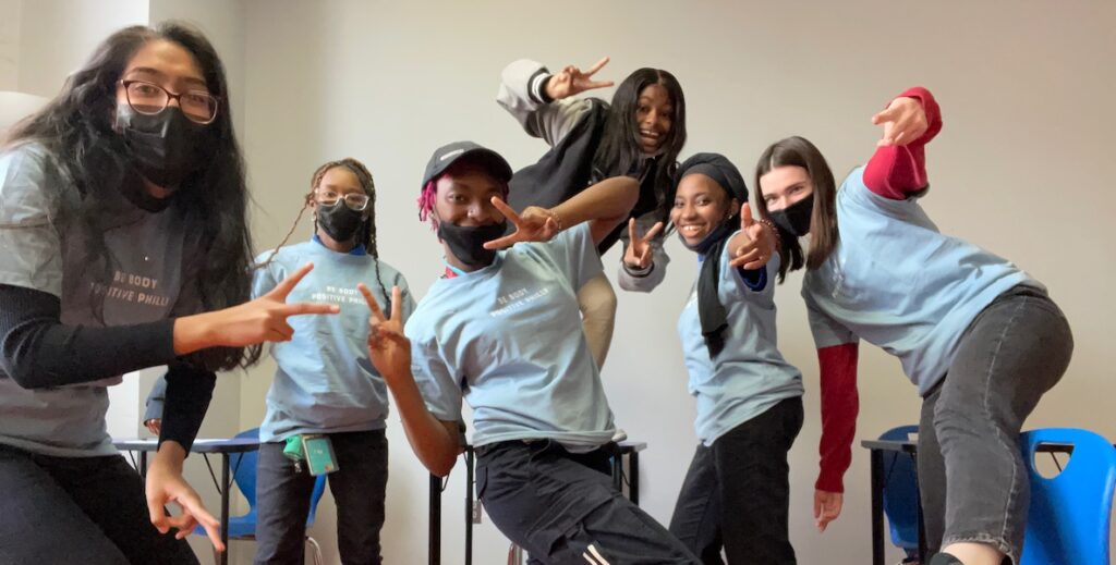 Six young women, all but one in light blue t-shirts and jeans, pose for the camera. One student stands on a chair behind the others, wearing a black varisty jacket. They are flashing peace and other positive finger signs.