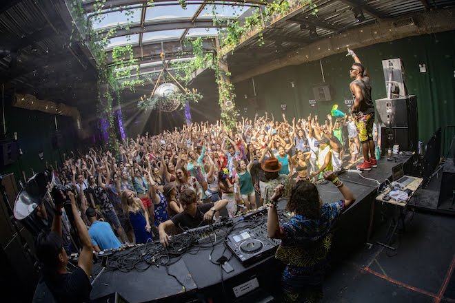 People wearing colorful clothing and headphones gather outside, dancing, before a stage where a DJ stands before a turntable and another person stands in front of the crowd, hand raised, during a Daybreaker alcohol-free rave.