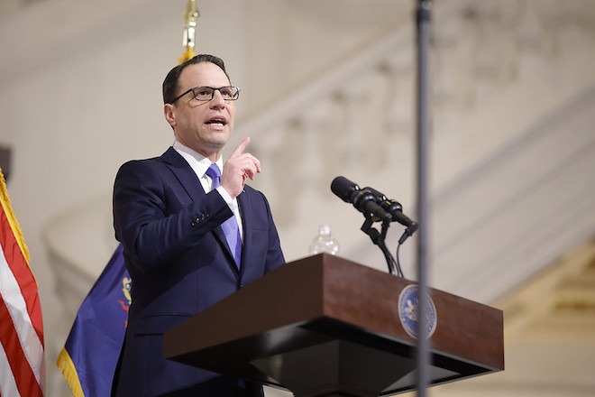 PA Governor Josh Shapiro a White man with short brown hair, glasses, wearing a blue suit, stands at a podium in front of the US flag, pointing a finger and giving his budget address.