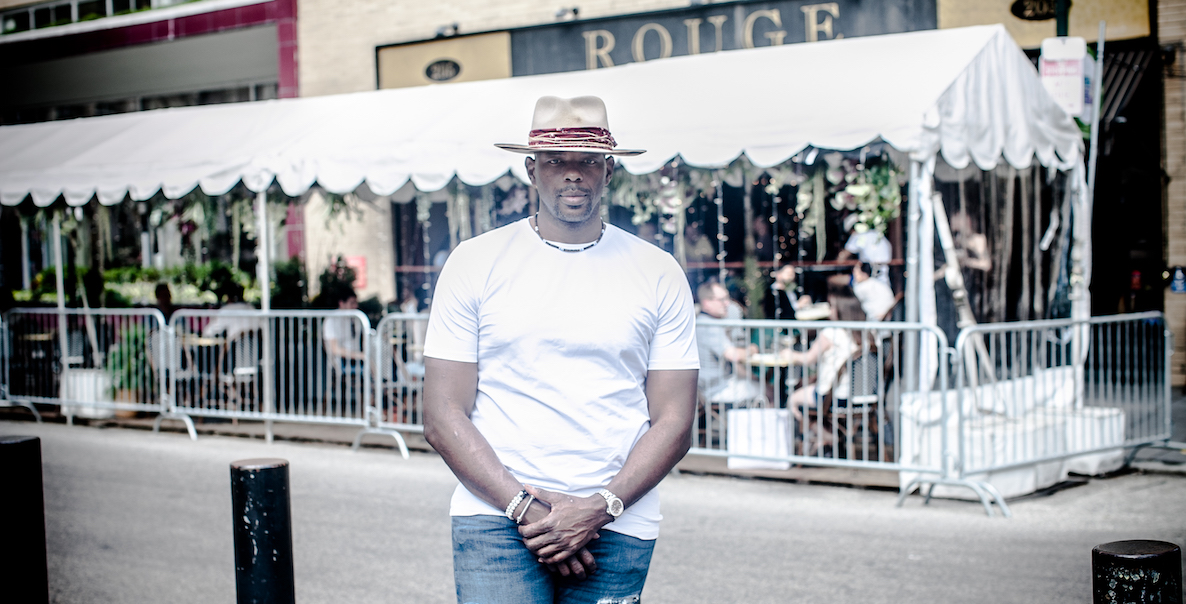 Marc Jackson, former NBA player and now 76ers TV analyst, stands across 18th Street in Philadelphia from Rouge, an indoor-outdoor bistro. He is a Black man wearing a wide-brimmed hat, white t-shirt, ripped jeans.