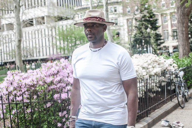 Marc Jackson, former NBA player and now 76ers TV analyst, azalea bushes in Rittenhouse Square. He is a Black man wearing a wide-brimmed hat, white t-shirt, ripped jeans.