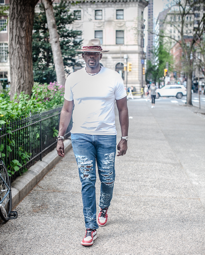 Marc Jackson, former NBA player and now 76ers TV analyst, walks along the sidewalk of Rittenhouse Square. He is a Black man wearing a wide-brimmed hat, white t-shirt, ripped jeans and Jordans.