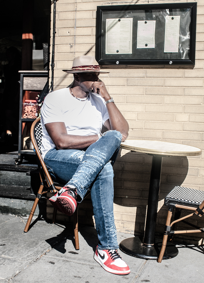 Marc Jackson, former NBA player and now 76ers TV analyst, sits outside of Rouge cafe in a chair on the sidewalk, leaning his elbow on a cafe table. He is a Black man in profile, with a wide-brimmed hat, white t-shirt, ripped jeans and Jordans, legs crossed.
