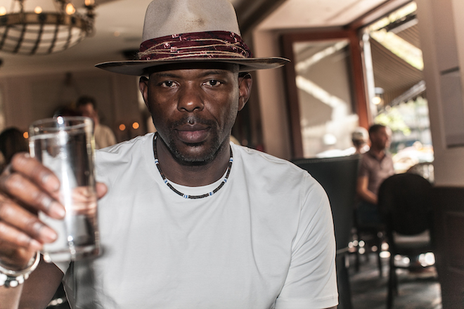 Marc Jackson, former NBA player and now 76ers TV analyst, sits in Rouge cafe holding up a glass of water. He is a Black man in a wide-brimmed hat, white t-shirt and necklace with a serious expression on his face.